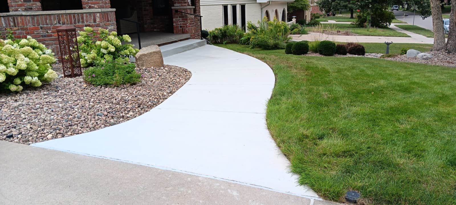 A curved concrete walkway running between a stone planting bed and a lawn toward a brick porch.